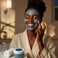 Woman applying our blue tea tree facial mask with a jar of mask and towel on a table in a cozy room.